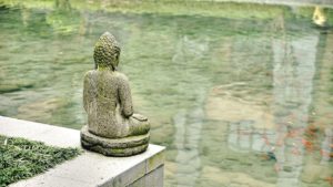 Stone Buddha statue seated overlooking still water. Buddha statue