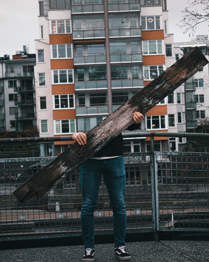 Man holding a plank of wood, but above the level of the plank he is missing - his face and body having disappeared from the photograph.